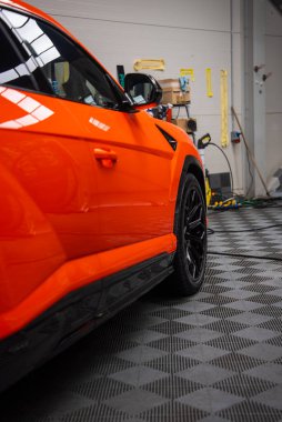 Parlak turuncu Lamborghini Urus SE bir atölyede oturur, parlak boya tavan ışıklarını, siyah alaşımlı tekerleği ve yan eteği yansıtır çerçeve, araçlar ve vites çizgisi gri.