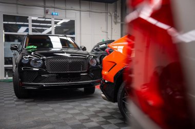 Kapalı mekandaki atölye sahnesinde turuncu Lamborghini Urus SE, parlak siyah bir Bentley Bentayga 'nın yanına park etmiş. Önünde kırmızı bir araba var..