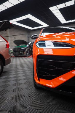 Yakından bakınca turuncu Lamborghini Urus SE bir atölyede, LED far, heykel tampon, parlak siyah ızgara, kapüşonlu ve kapağı açık arabalar görünüyor..