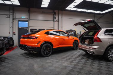 Turuncu bir Lamborghini Urus SE gri kareli bir zeminde, parlak panel aydınlatmasının altında, duvarda ve yakınlarda gümüş renkli bir SUV de dahil olmak üzere araçlarla duruyor..