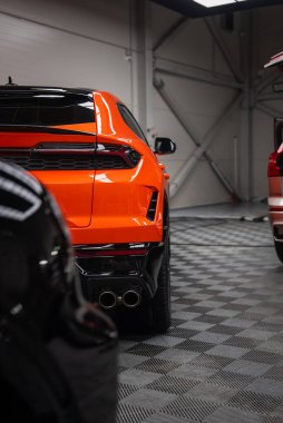 Turuncu Lamborghini Urus SE gri kareli zeminde, arka üç çeyrekte otururken arka tarafta egzozlar, keskin stop lambaları, yontulmuş tamponlar, yan yana park edilmiş arabalar görülüyor..