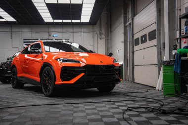 Turuncu bir Lamborghini Urus SE, Y şeklindeki LED farları, siyah aero aksanları, büyük siyah tekerlekleri, detay araçları ve sarmalanmış güç kablosu olan iyi ışıklandırılmış bir atölyede duruyor..