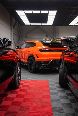 Açık turuncu bir Lamborghini Urus SE koyu renkli ciplerin arasına park edilmiştir. Dörtlü egzozlar, siyah difüzyon ve keskin stop lambaları endüstriyel tavan lambaları altında göze çarpıyor..