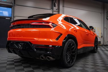 Turuncu Lamborghini Urus SE bir kapalı servis koyunda, alçak arka üç çeyrek görünüm oyulmuş stop lambaları, dörtlü egzoz uçları, siyah difüzör ve büyük siyah alaşımlı tekerlekler gösteriyor.