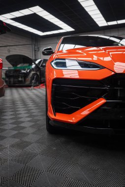 Yakından bakıldığında turuncu bir Lamborghini Urus SE ön tarafı, LED far, büyük siyah ızgara ve servis altında arabaları ve floresan panelleri olan bir atölyede keskin yüzgeçler görülüyor..