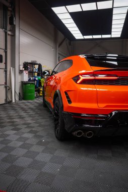 Detaylı bir bölmede turuncu Lamborghini Urus SE, alçak arka üç çeyrek görünüm dörtlü egzoz, açısal stop lambaları, parlak siyah difüzyon, panel ışıklandırması ve kareli zemin.