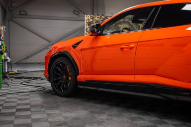 Parlak turuncu Lamborghini Urus SE bir atölye, ön sol çeyrek panel, kapı ve turuncu kalibre odaklı siyah çoklu konuşma tekerleği, parlak bitiş.
