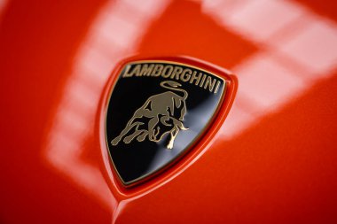 Yakın plan Lamborghini armasını kapalı bir atölyede turuncu bir kaputta gösteriyor. Hafifçe yukarıdaki ışıklar boyayı yansıtıyor, ayrıntıları ve zanaatı vurguluyor..