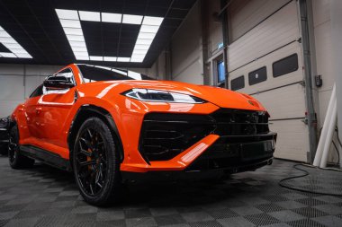 Alçak açı turuncu Lamborghini Urus SE 'yi bir atölyede, LED farlar, siyah aero, çok konuşan tekerlekler, turuncu kalibreler, aydınlatma paneli, garaj kapıları.