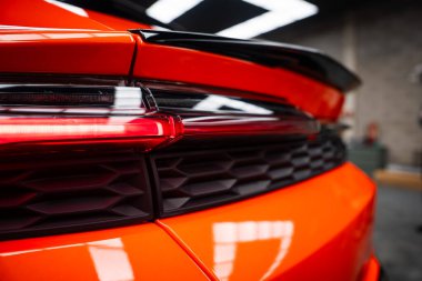 Atölyede turuncu bir Lamborghini Urus SE 'nin yakın görüntüsü, arka lambaya odaklanma, siyah bal peteği menfezi, ve parlak boya üzerine yansıyan parlak ışık ile oyulmuş spoiler.