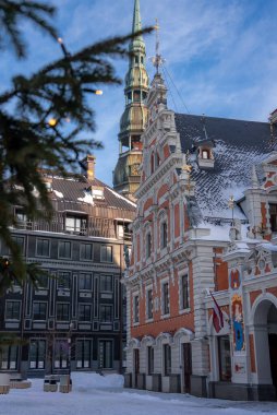 Kara Kafalar Evi Letonya Riga 'da kar kaldırım taşlarının tozunu alırken duruyor. Aziz Peter Kilisesi arkasında yükseliyor. Letonya bayrağı ve peri ışıkları Noel öğleden sonra.