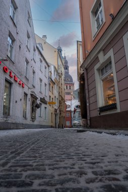 Riga, Letonya 'da dar bir kaldırımtaşlı sokak Riga Katedrali kulesine ve bakır kubbeye çıkıyor. Isıtıcı dükkanı penceresinin ışığı parlıyor. Düşük açı, buzlu taşları ve yüksek ortaçağ binalarını gösteriyor..