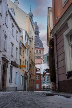 Riga Eski Kasabası 'ndaki dar bir kaldırım taşı yolu Riga Katedrali' ne çıkıyor. Kar kenarlara ve silindirlere dayanır. Letonya bayrakları ve işaretleri beliriyor. Yumuşak, serin gün ışığı sakin bir ruh hali yaratır..