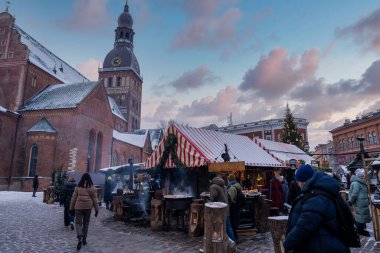 Riga Katedrali ve saat kulesi Letonya 'nın Riga kentindeki bir Noel pazarında yükseliyor. Kar, dumanı tüten yemek tezgahları, çizgili çatılar ve çelenkler akşam manzarası hazırlıyor..