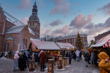 Riga, Letonya, Dome Square Riga Katedrali ve saat kulesini Noel pazarı olarak gösteriyor. Tezgahlar, sicim ışıkları ve alacakaranlıkta karla kaplı uzun bir ağaç..