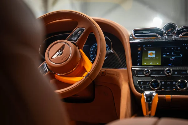 Yakın iç görünüş kahverengi deri tekerlek, kanatlı B, Breitling saati, karbon fiber tire, MIX FM 102.7 ekranda, aydınlatma göstergeleri, turuncu aksanlar, showroom ışıklandırması gösteriyor..