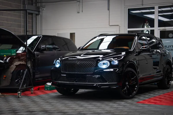 Parlak siyah Bentley Bentayga Speed, parlak atölye ışıkları altında, matris ızgarasını, oval ikiz farları, siyah alaşımlı tekerlekleri, aletleri ve kareli paspasları gösteren kapalı bir yerde oturuyor..