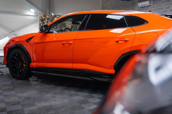 Yakından bakıldığında turuncu bir Lamborghini Urus SE 'nin parlak boya, siyah tekerlekler, turuncu kalibreler, kareli döşemeler ve detaylı ekipmanlarla modern bir atölyeye park edildiğini görüyoruz..