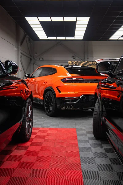 Açık turuncu bir Lamborghini Urus SE koyu renkli ciplerin arasına park edilmiştir. Dörtlü egzozlar, siyah difüzyon ve keskin stop lambaları endüstriyel tavan lambaları altında göze çarpıyor..