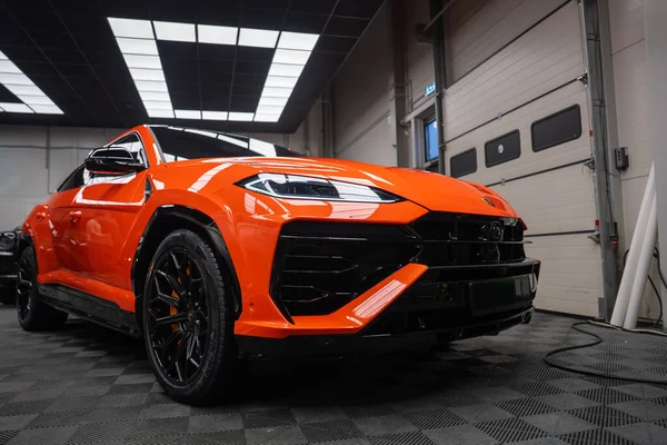 Alçak açı turuncu Lamborghini Urus SE 'yi bir atölyede, LED farlar, siyah aero, çok konuşan tekerlekler, turuncu kalibreler, aydınlatma paneli, garaj kapıları.