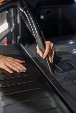 Bir kişi, ayrıntılı bölmede dar bir çatlak olan bir arabanın bagaj dikişini vakumluyor, görev aydınlatma mevcut, koyu renk süsleme ve leopar desenli tırnak sanatı görünür..