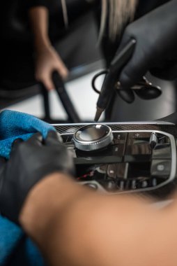 İki eldivenli el modern bir araba konsolunu fırça, hava aracı, vakum ve mavi bir mikrofiber bez kullanarak temizliyor. Sığ alan derinliği ve yansıtıcı yüzeyleri olan kapalı bir alan..