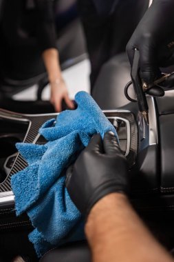 İki eldivenli el modern bir araba merkezi konsolunu, vites seçici ve karbon fiber süslemeli, yumuşak ışıklandırmalı ve dar çerçeveli parlak mavi bir bez kullanarak temizler..