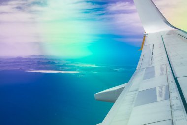 Bir uçak penceresinden gelen hava görüntüsü engebeli arazi şekilleri, beyaz kum girişleri, sis, prizmatik ışık parlamaları ve yüksek irtifa bulutları olan derin mavi bir kıyı şeridi üzerindeki kanat ve motoru gösteriyor..