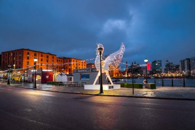 Liverpool Royal Albert rıhtımının rıhtımına ışık tutan tuğla depoları. Ciğer Kuşu heykeli Albert Dock ve Barnacle 'ın tabelalarının yanında duruyor. Suyun üzerinde bir dönme dolap parlıyor, ıslak kaldırım taşları kış yağmurunda parlıyor..