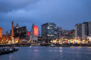 Alacakaranlıkta Liverpool 'daki Royal Albert Rıhtımı, Kraliyet Karaciğer Binası' nın saat kuleleri ve ilerideki uzun kırmızı ışıklı kule, Noel ışıkları ve market tezgahları rıhtımda..