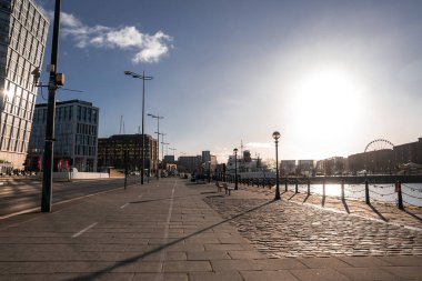 Liverpool 'daki güneşli gezinti güvertesinde açık bir kış gününde sol taraftaki ofisler, demirli gemi ve zincirler rıhtım sağ, ufuk çizgisi ve yankı tekerleği görünüyor..