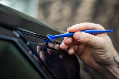 Bir teknisyen, sığ bir alan derinliği ve kapalı bir aydınlatma ile, bir bayilik servis bölümünde parlak siyah bir araba penceresinin dar bir lastik mührünü kaldırmak için mavi plastik bir budama aracı kullanır..