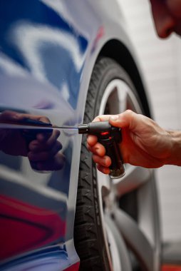 Bir teknisyen, parlak mavi bir arabanın kapısındaki hafif ışıklandırmalı, profesyonel bir bakım hangarındaki spor alaşımlı tekerleğin üzerindeki küçük bir çiziği onarmak için küçük bir meşale kullanır..