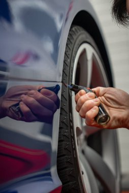 Bir teknisyen, kapalı alanda parlak mavi bir arabanın arka tekerlek kemerinin yanındaki plastik süslemeyi ısıtır ve düzleştirir. Boya, hafif ışık ve lastik izleri görünür..