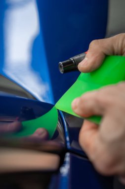 Eller, ısı tabancası kullanarak mavi lüks araba paneline parlak yeşil bir örtü uygular. Bir krupiye dükkanında kapalı alan, sığ alan derinliği ve yansımalar hassas çalışmayı vurgular..