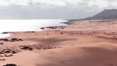 Hava kumulları, Atlantik sörfü, lav kayaları ve Fuerteventura, Kanarya Adaları 'ndaki düz tepeli kütle. Dar kıyı yolu, küçük arabalar ve plaja gidenler güneşli bulutların altında ölçek gösteriyor..