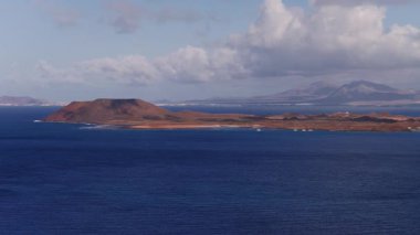 Gündüz kuşağı Fuerteventura 'nın Lobos adasını, pas kumullarını, Atlantik dalgalarını, yelkenlileri ve dalgalı bulutların altındaki Lanzarote dağlarını gösteriyor..