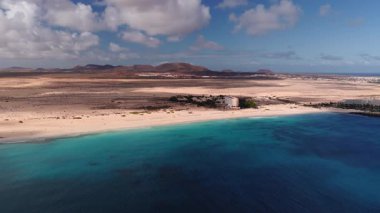Hava 4K görüntüsü, beyaz kumsal, turkuaz çivit denizi, kurak kum tepeleri, volkanik tepeler, alçak katlı oteller, güneşlikler, hafif dalgalar, rüzgar türbinleri, Lanzarote ufku gösteriyor..