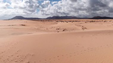 Fuerteventura 'daki Corralejo kumullarının alçak bulutların altındaki hava manzarası. Dalgalı kum, ovalama, ayak izleri, park etmiş arabalar, küçük aylaklar ve düz tepeli bir koni görülebilir..