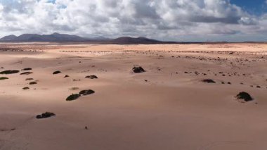 Hava taraması Fuerteventura 'da soluk kum tepeleri, düz çöl ovaları ve volkanik siluetler gösteriyor. Rüzgar türbinleri, park etmiş arabalar, küçük insanlar ve güçlü güneş gölgeleri..