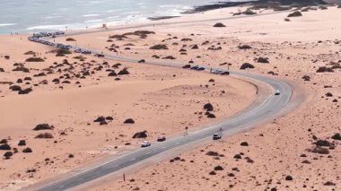 Hava 4K sahnesi Fuerteventura, Kanarya Adaları 'ndaki turuncu kum tepecikleri boyunca uzanan, hareket eden araçlar, sörf, temizlik ve küçük figürlerle kıvrımlı bir kıyı yolu gösteriyor.
