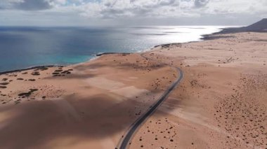 Hava aracı görüntüsü, Fuerteventura 'daki kayalık bir sahile ve volkanik koniye giden iki şeritli bir yol gösteriyor. Arabalar, dalgalar, bulut gölgeleri ve yumuşak ışık..