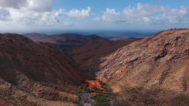 Havadan gün ışığı süzülüyor ve tavada paslı tepeler, katmanlı kayalar, yeşil yamalar ve yuvarlanan volkanik tepeler ve denizin soluk bir adası ile küçük beyaz bir yapı görünüyor..