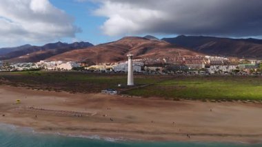 Fuerteventura, Morro Jable ve Jandia 'nın arkasındaki beyaz deniz fenerinin havadan görünüşü, turkuaz sörf, kum tepesi şeridi, oteller, güneş panelleri ve bulut gölgeleri.