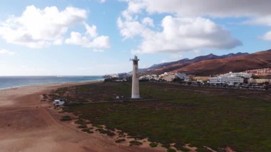 Fuerteventura 'daki Morro Jable deniz fenerinin havadan görünüşü. Yürüyüşçüler kumlu sahil boyunca hareket eder, sörf turları, tatil köyleri ve volkanik tepeler arkasında, Atlantik solda..