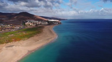Hava görüntülerine göre Fuerteventura hilal plajı, turkuaz deniz suyu, kumlu sahil, sahil beldesi, volkanik tepeler, kum tepeleri, ufukta Lanzarote, hafif dalgalar..