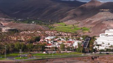 Beyaz otel, villa, metal heykel, golf sahası, volkanik tepeler, dağ yolu, arabalar, otobüsler ve yayalar ile Fuerteventura tatil köyünün hava sahnesi..