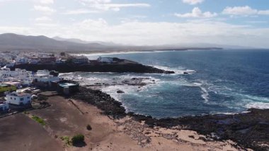 Fuerteventura 'daki siyah volkanik kayalıklardaki bembeyaz evlerin hava manzarası. Turkuaz sörf kayalıkların üzerinde kırılır, köpük lav kıyılarıyla buluşur, uzun sahil, puslu dağlar.