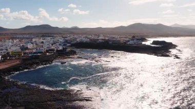 Hava manzarası Fuerteventura 'daki karanlık volkanik kayaların üzerinde beyaz boyalı bir sahil köyü gösteriyor. Korunaklı bir körfez, dalgalar, gelgit havuzları ve ılık gün ışığı altında alçak tepeler var..