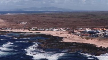 Fuerteventura 'nın havadan görünüşü, Kanarya Adaları. Atlantik dalgaları volkanik kayaları ve kumlu plajları vurur. Resort 'lar kıyı boyunca uzanıyor. Lanzarote ufukta oturuyor. Sahile gidenler yürür.